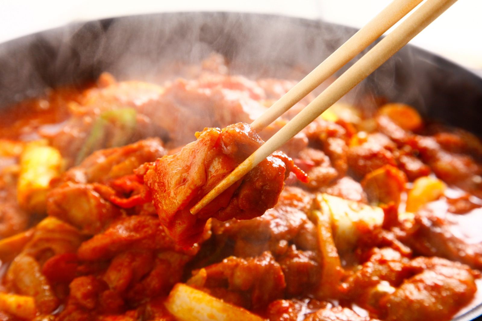 A close-up of dakgalbi cooking in a black pan, chicken glazed in red sauce with steam rising