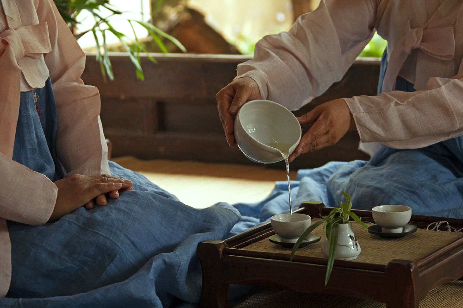 Two people in hanbok performing a traditional Korean tea ceremony, pouring green tea at a low wooden table