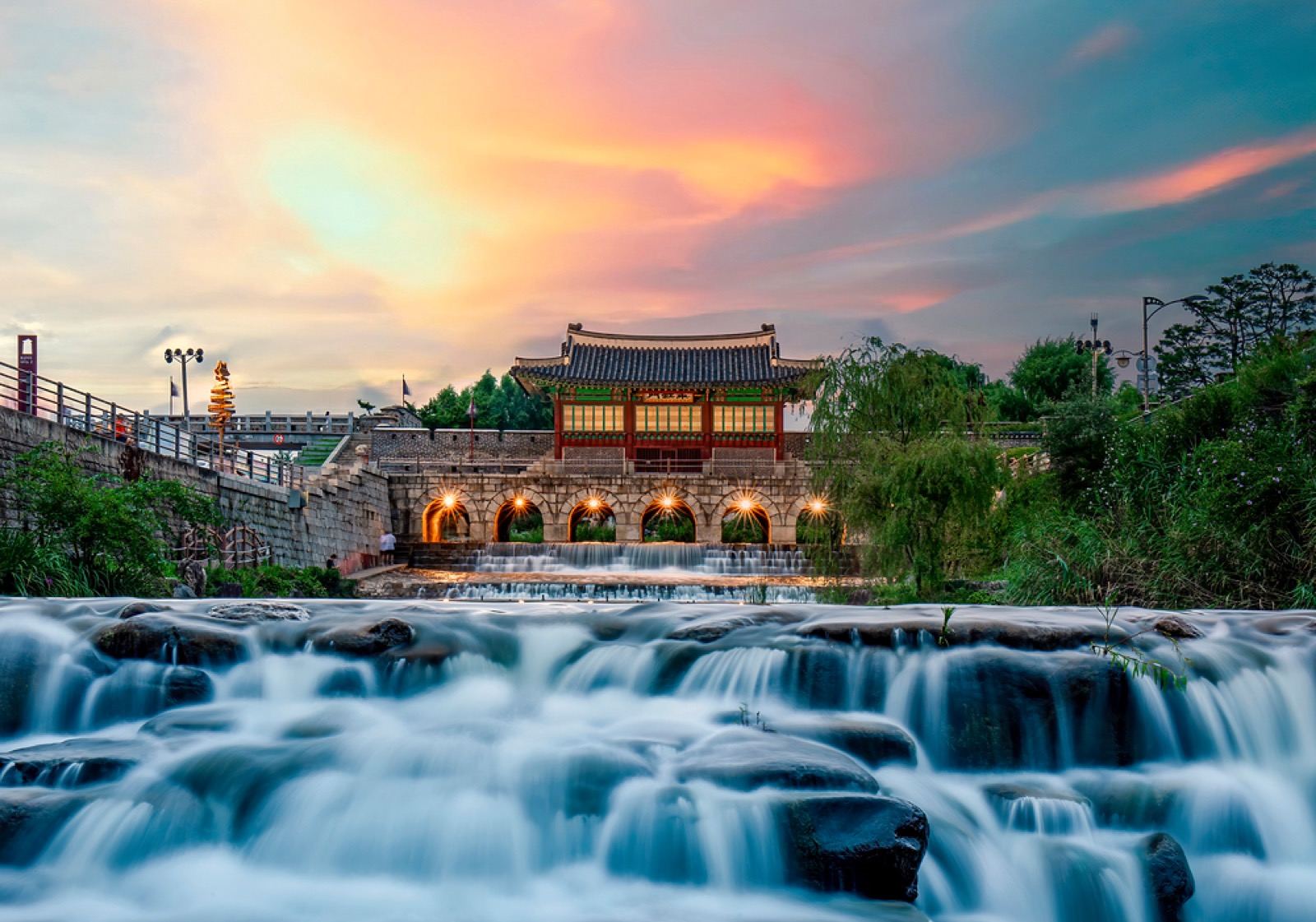 Hwahongmun water gate lit at sunset, its arches glowing above a long-exposure waterfall on the stream below