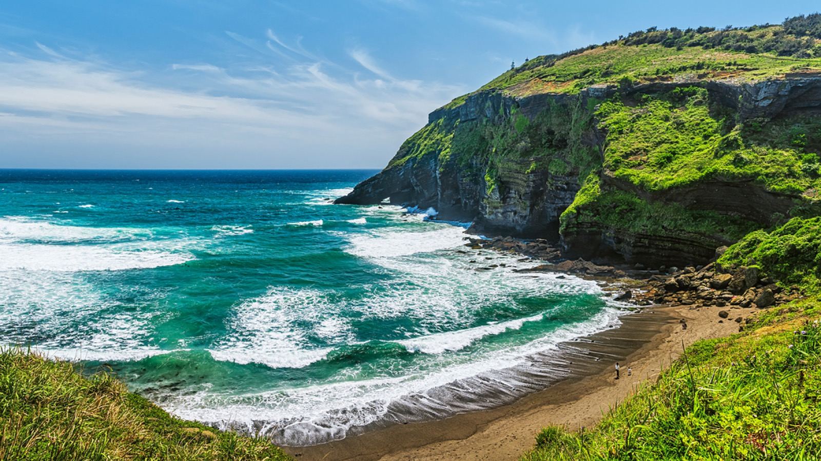 A green coastal cliff above a curving beach and turquoise water on Udo island, off Jeju