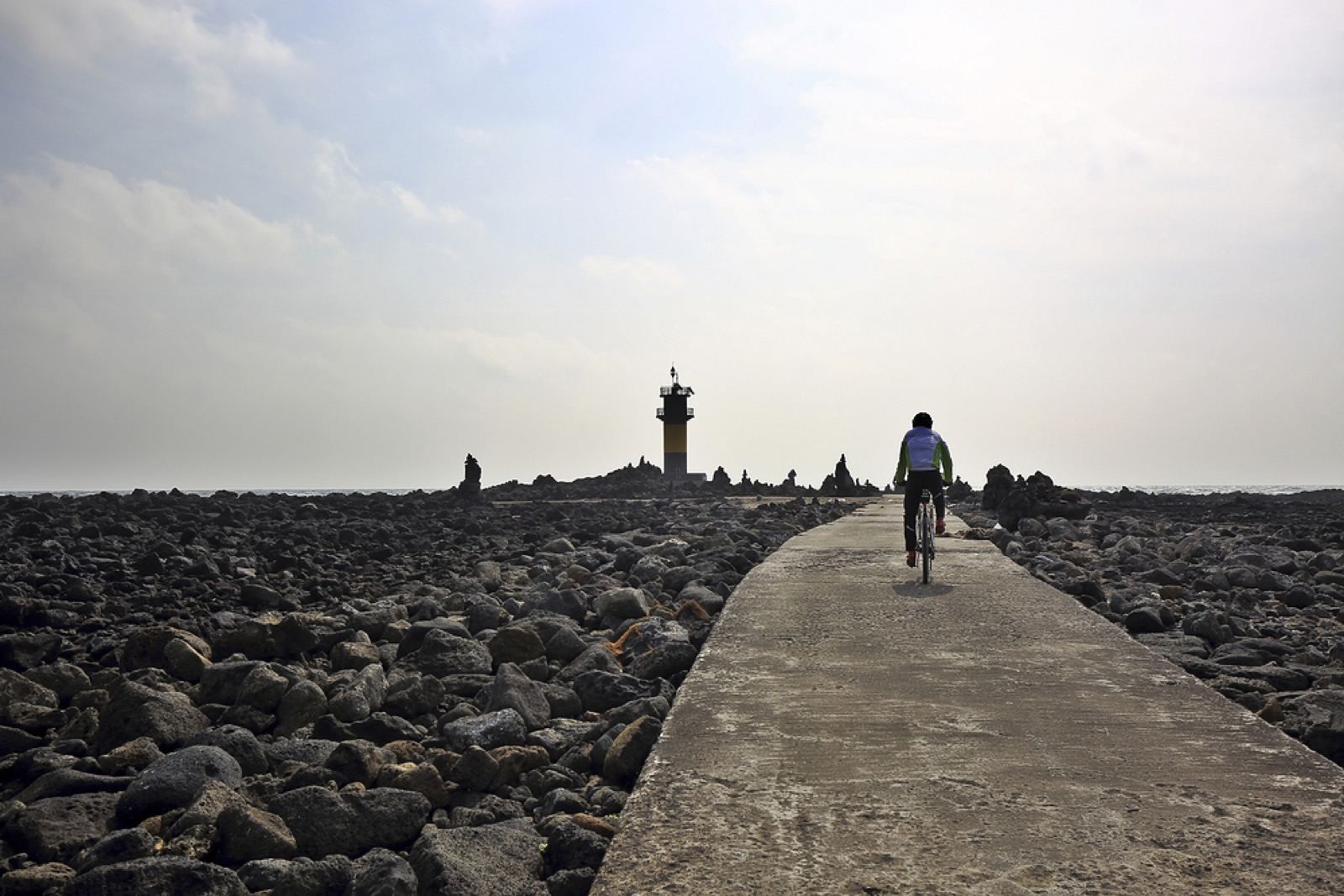 A cyclist riding a stone causeway toward a small lighthouse across a field of black volcanic rock on Udo